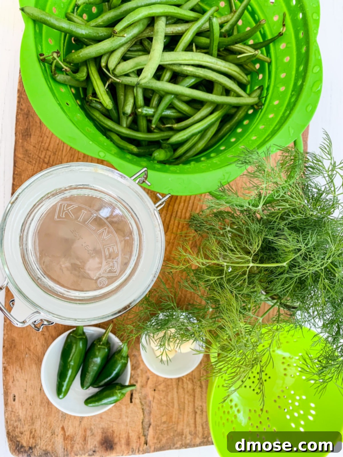 A cutting board with a green colander filled with green beans and pickling ingredients from a top-down view.