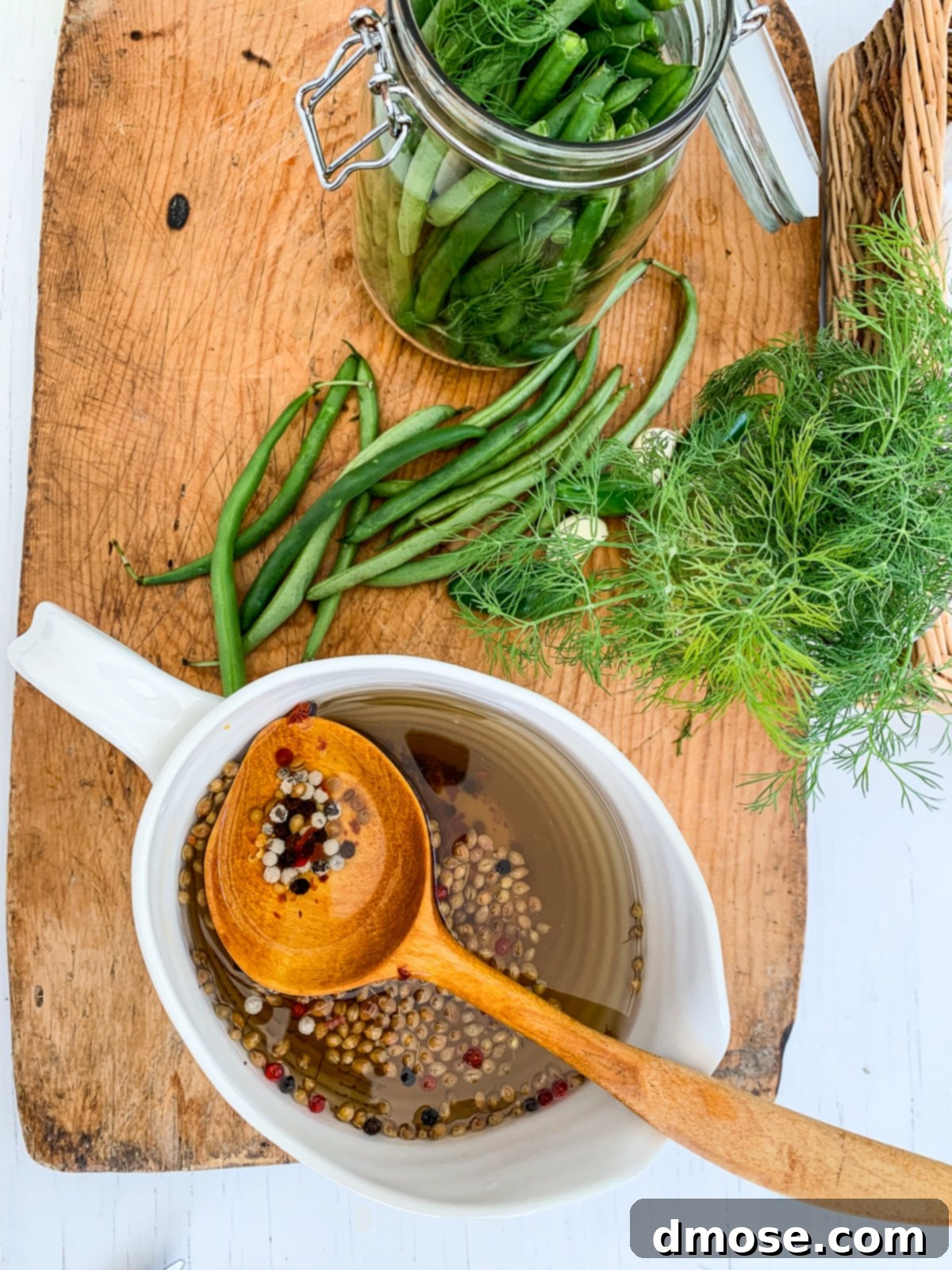 A cutting board with all the ingredients arranged for making green bean pickles, including fresh dill, garlic, and red pepper flakes.