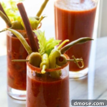 A pitcher of bloody marys with two drinks garnished with olives, pepperoncinis, and beef sticks.