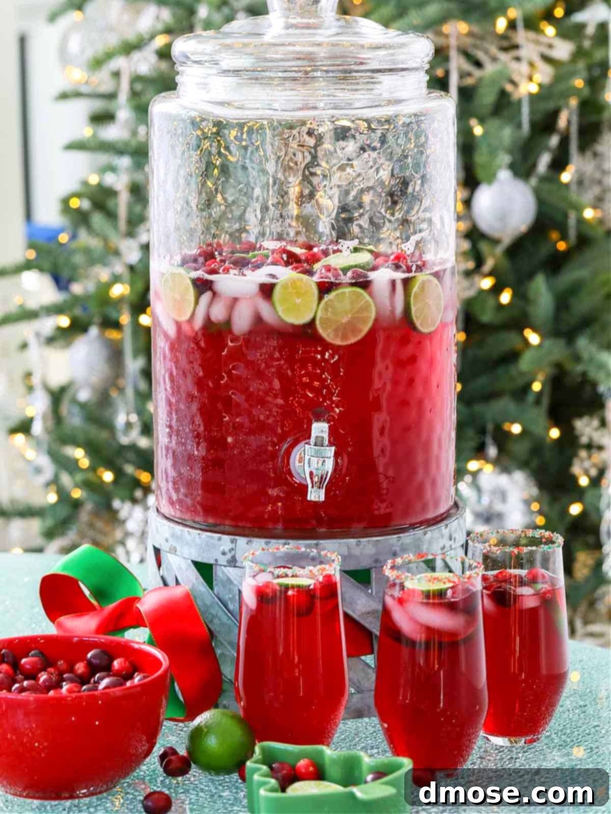 A large glass beverage dispenser filled with vibrant red non-alcoholic Christmas Punch, garnished with fresh cranberries and lime slices.