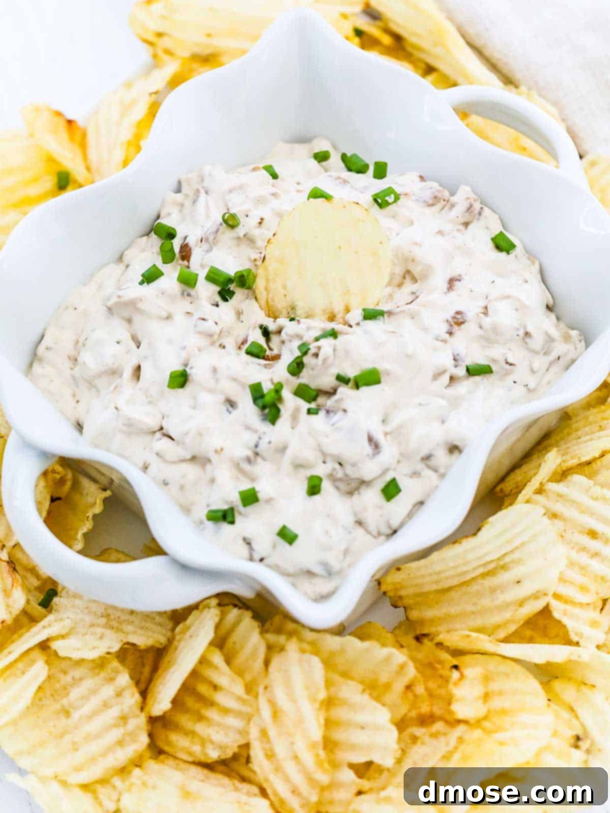 A serving dish filled with creamy French Onion Dip, garnished with fresh chives and a potato chip standing upright.