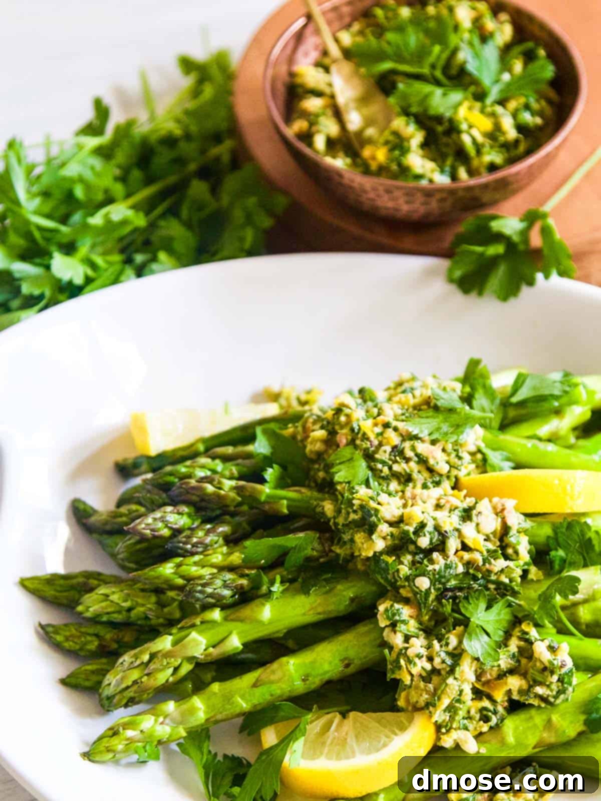 Grilled Asparagus spears generously topped with vibrant Gremolata and fresh lemon wedges on a white plate.