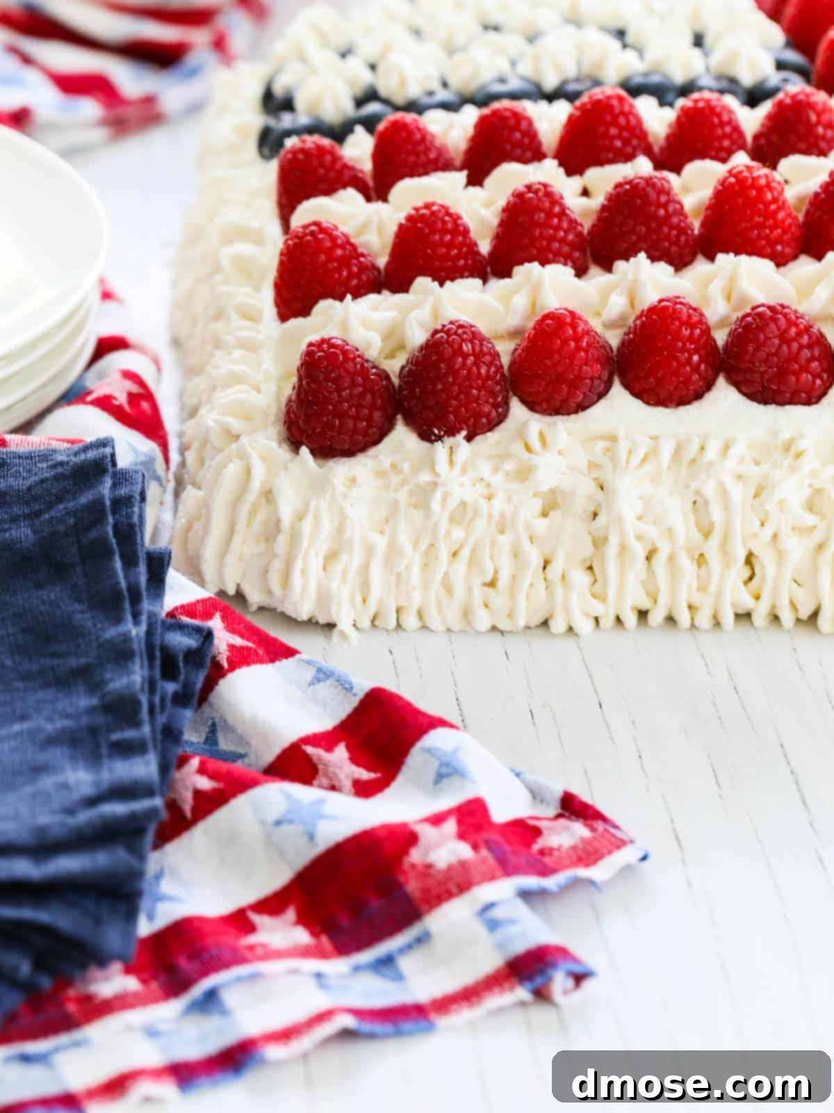 The corner of a white frosted American Flag Cake decorated with raspberries, with a patriotic towel nearby.