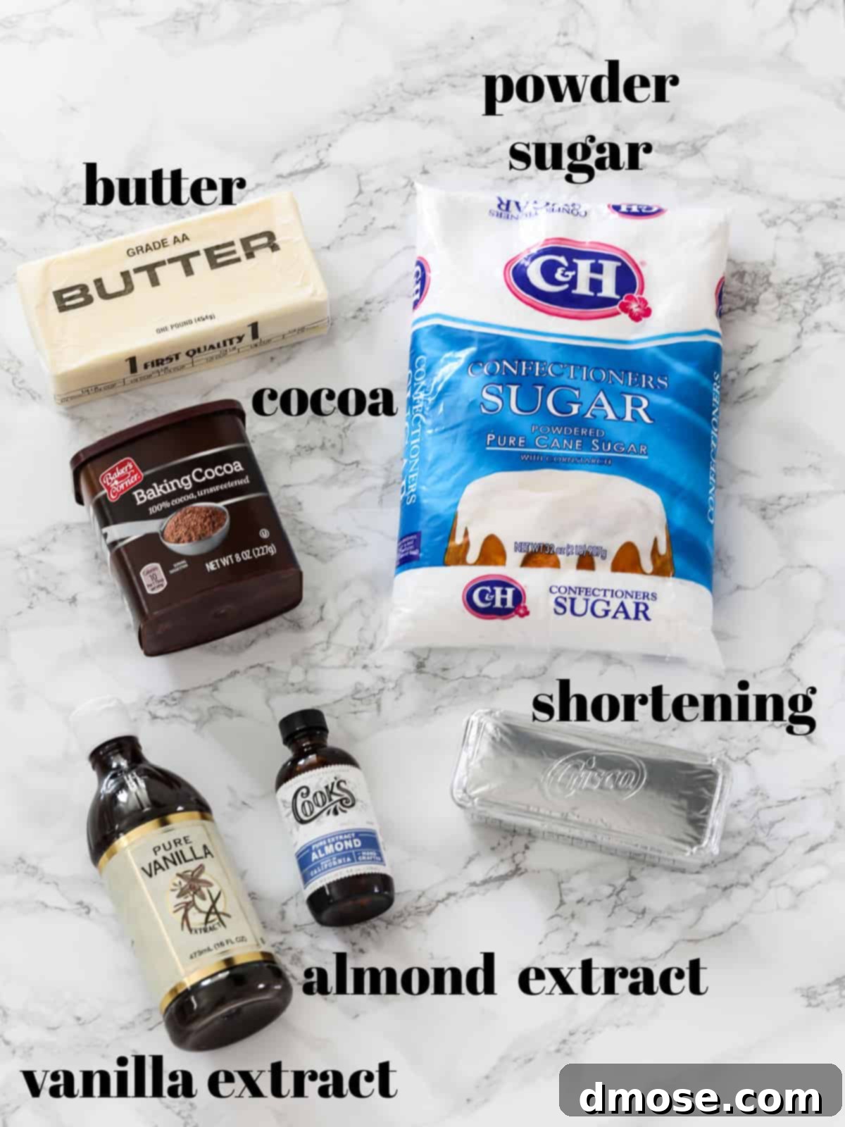 All the ingredients laid out on a table for making homemade chocolate frosting: butter, shortening, powdered sugar, cocoa powder, vanilla extract, almond extract, and hot water.