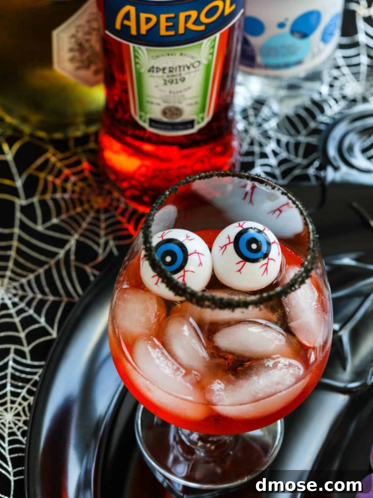 Wicked Aperol Potions 5 A close-up of a Halloween Aperol Cocktail featuring a black sugar rim, plastic eyeballs, and a vibrant orange hue, set against a spooky backdrop.