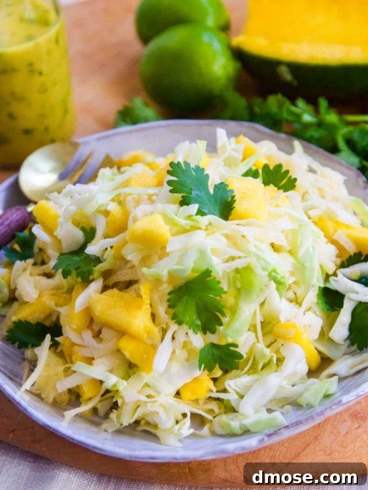 Mango slaw piled on a plate with a glass jar of dressing and limes in the background.