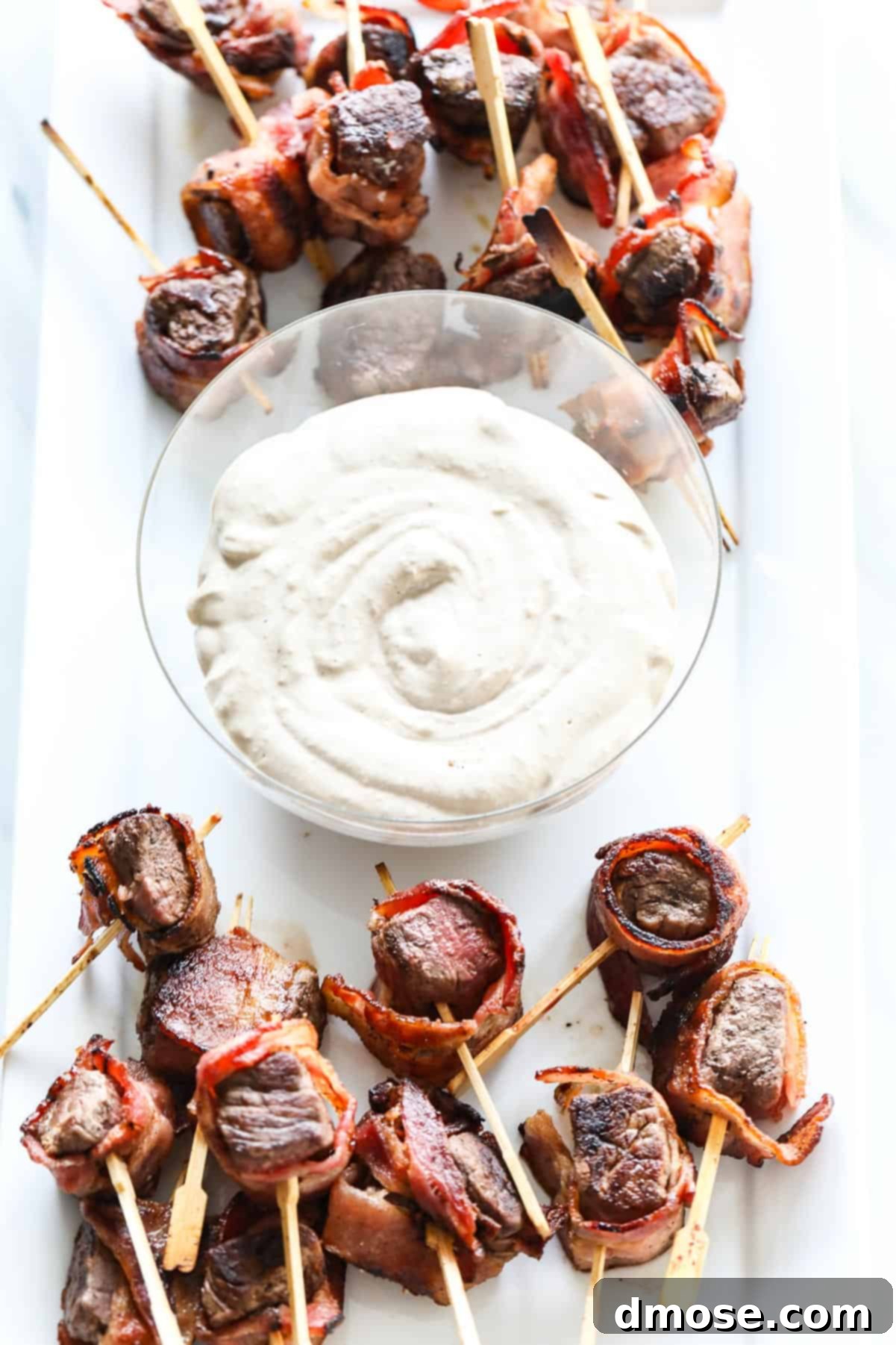 Bacon-wrapped steak appetizers on a platter with a bowl of creamy horseradish sauce.