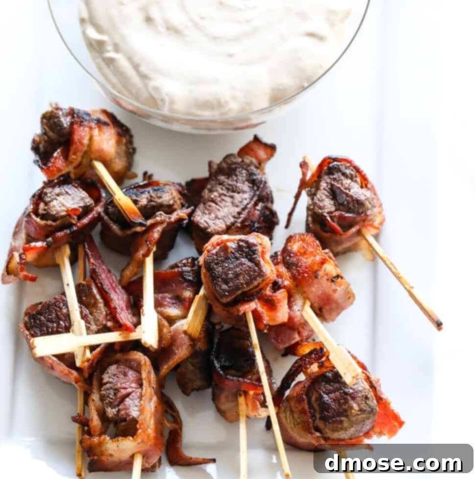 A glass bowl filled with horseradish dipping sauce with bacon wrapped filet mignon appetizers on toothpicks.