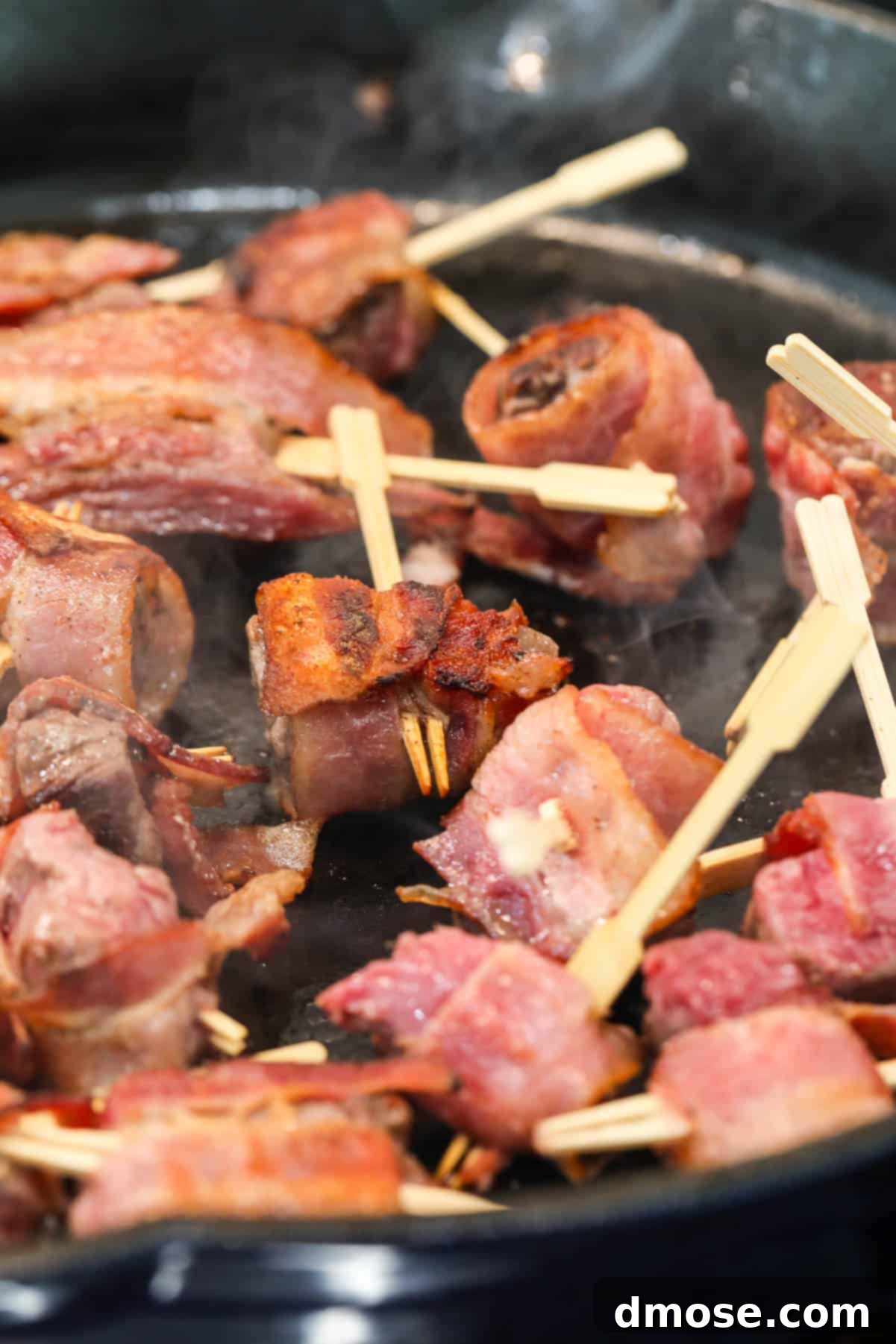 More bacon-wrapped steak appetizers searing in a cast iron pan, showing the rich color development.