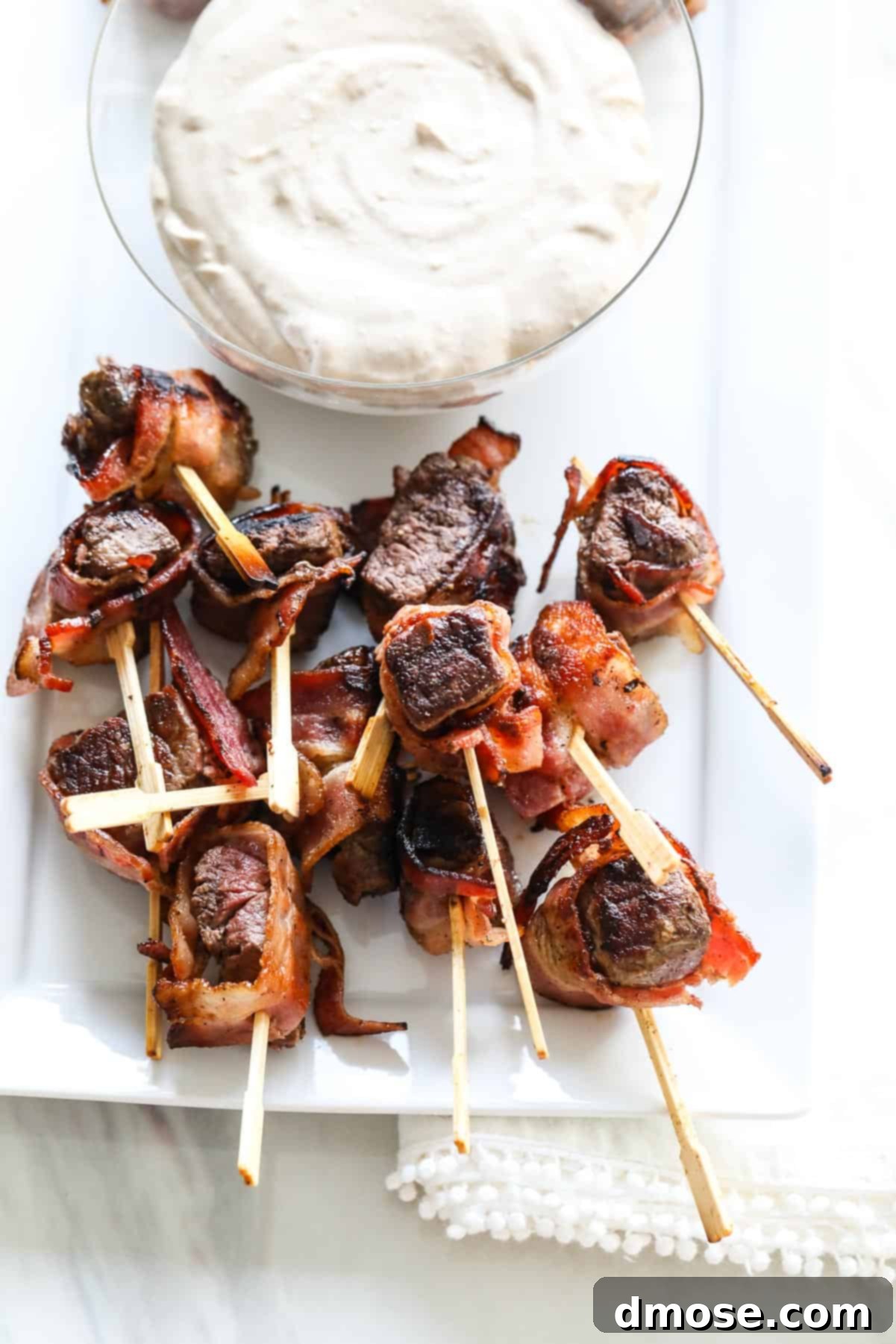 Platter of bacon-wrapped steak appetizers with a bowl of creamy horseradish sauce, garnished with fresh herbs.