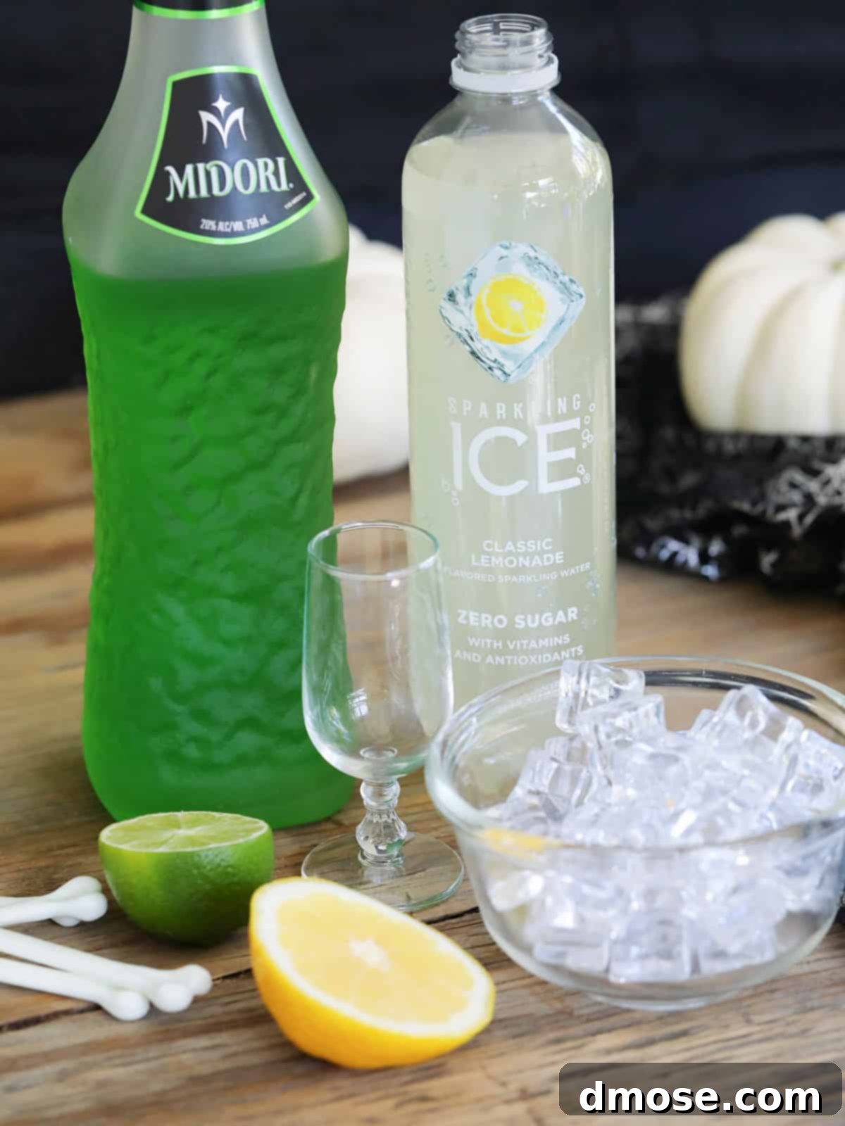 Mystic Melon Elixir 4 A collection of fresh ingredients for Midori sours laid out on a table, including Midori liqueur, ice, vibrant lemons, juicy limes, vodka, and sparkling water.