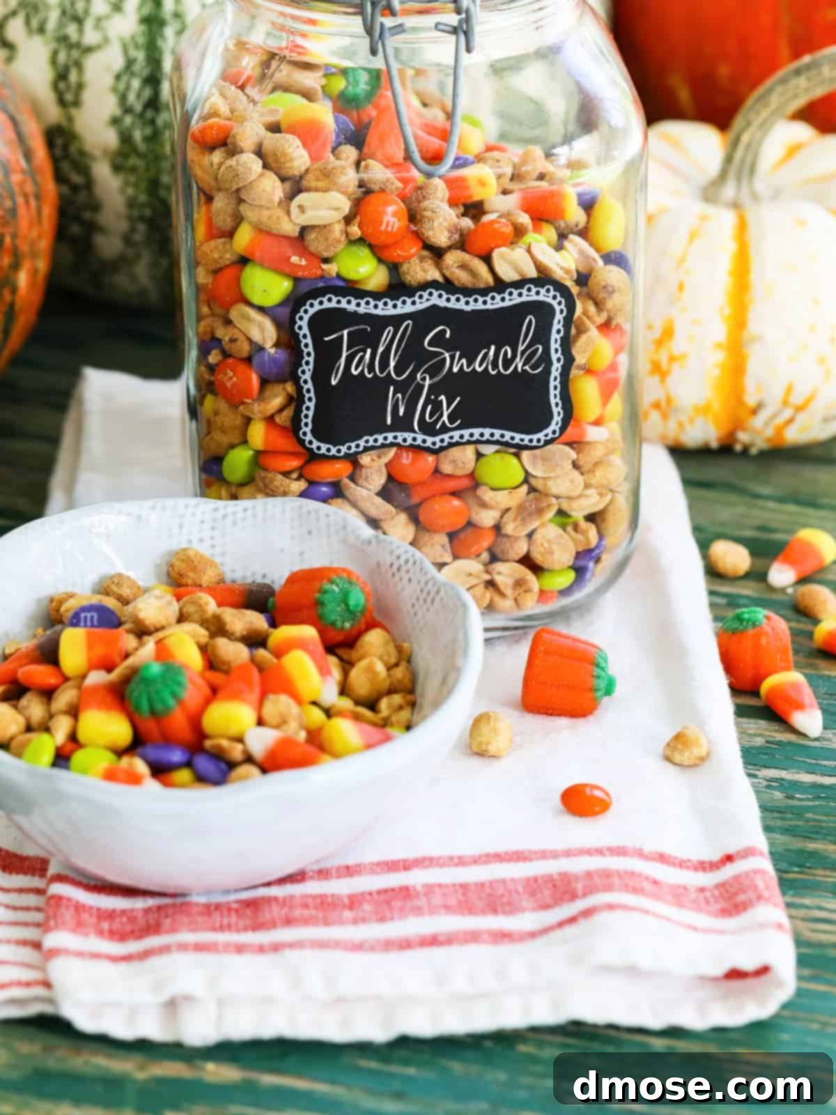 A large glass jar filled with a colorful Fall Snack Mix featuring candy corn and M&Ms, with a smaller bowl of the mix nearby.