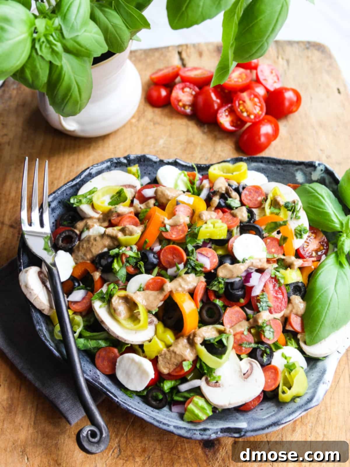 A vibrant Pizza Salad on a white ceramic plate, featuring mini pepperoni, black olives, chopped lettuce, tomatoes, and mozzarella balls, drizzled with dressing.