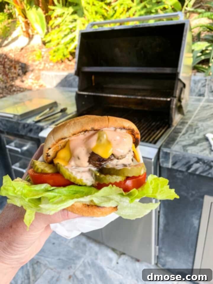 A person holding a messy, mouth-watering grilled hamburger wrapped in parchment paper, standing beside an open stainless steel gas grill, ready for a delicious game day meal.