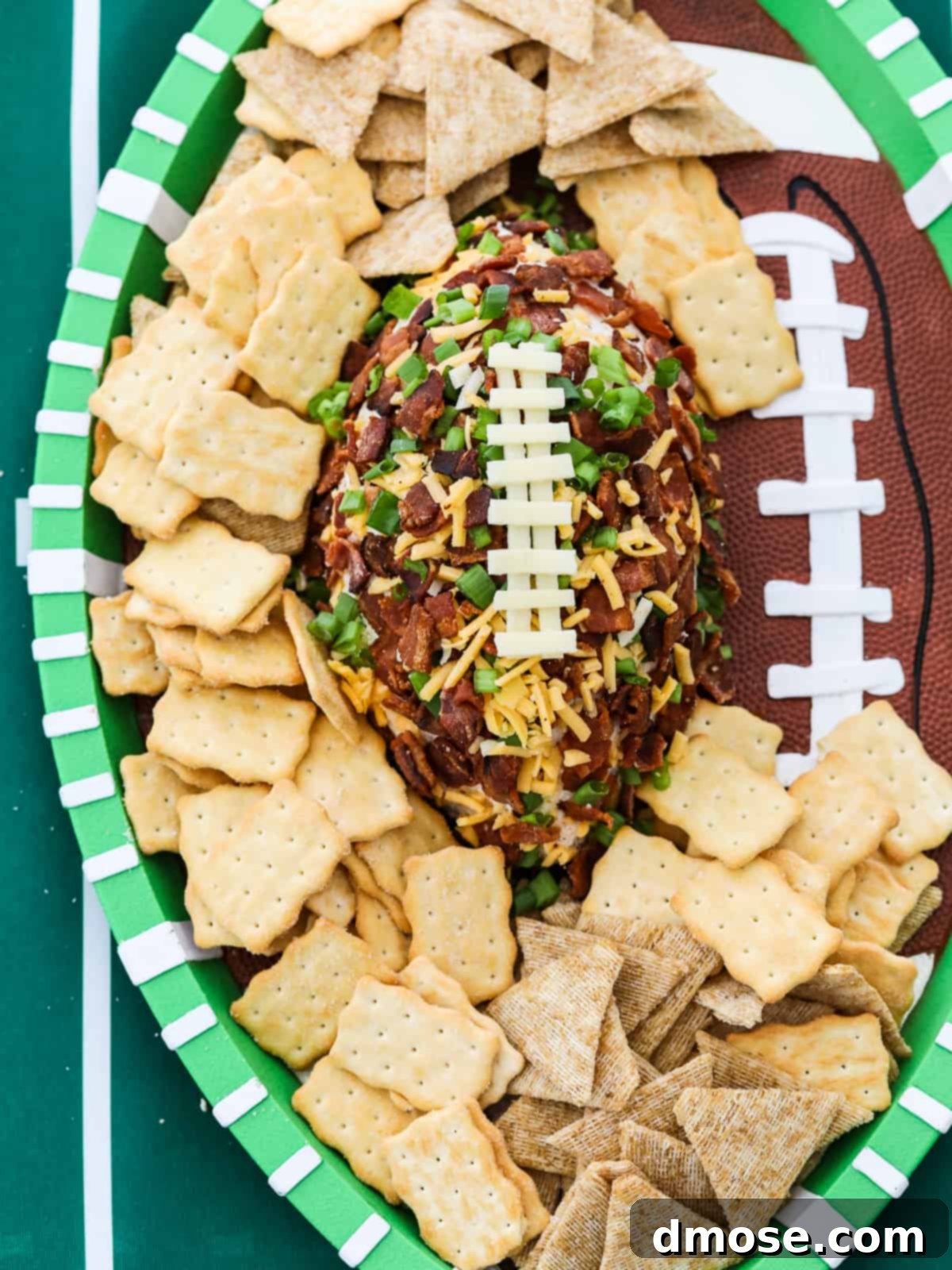 A football shaped cheese ball coated with crumbled bacon, green onions, and cheddar cheese and white cheese to look like laces on the football surrounded by crackers on a platter.