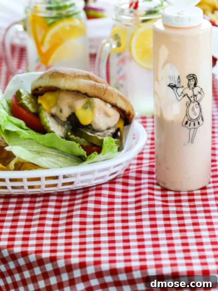 A messy grilled burger topped with lettuce, tomato, cheese, pickles and burger sauce in a basket with a bottle of burger sauce.